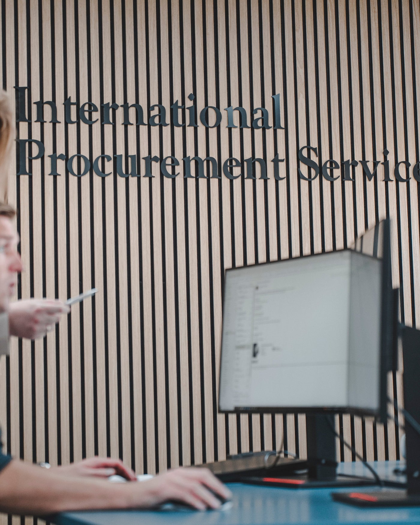 International Procurement Service employee sitting in front of a computer screen with logo in the background.